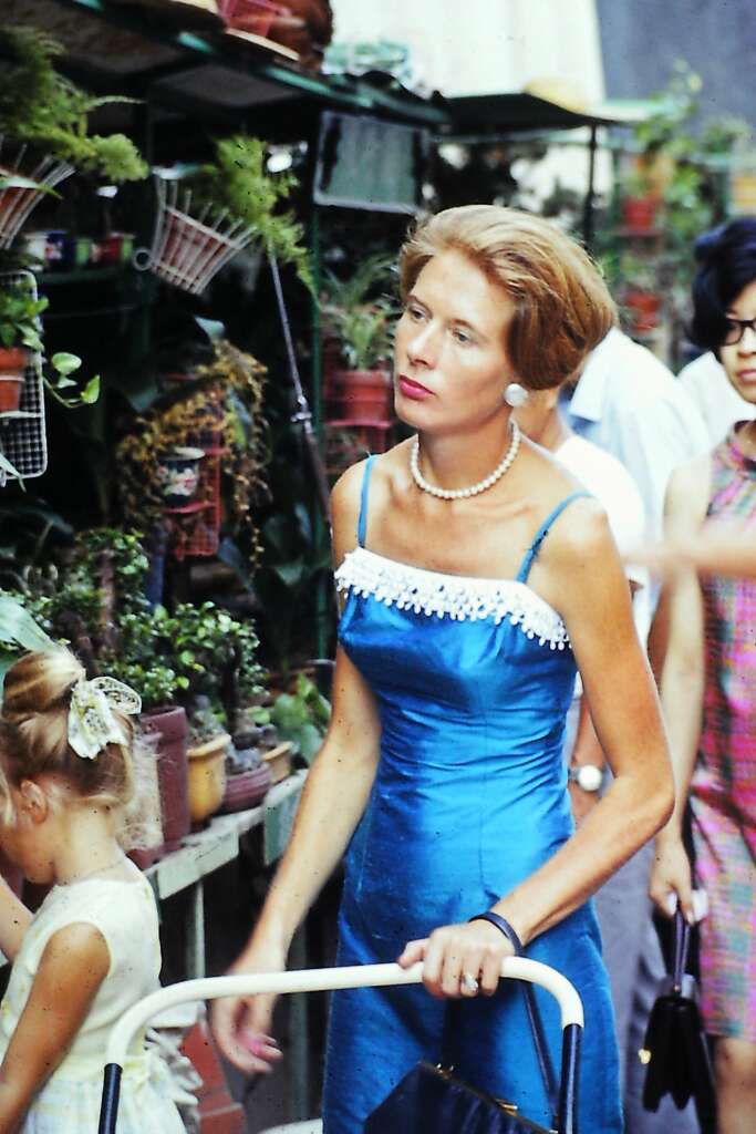 A woman with red hair, wearing a blue sleeveless dress with white trim and a pearl necklace, pushes a white frame overgrowth of potted plants. A young girl with blonde hair is beside her, and a woman with dark hair stands behind them, partially visible.