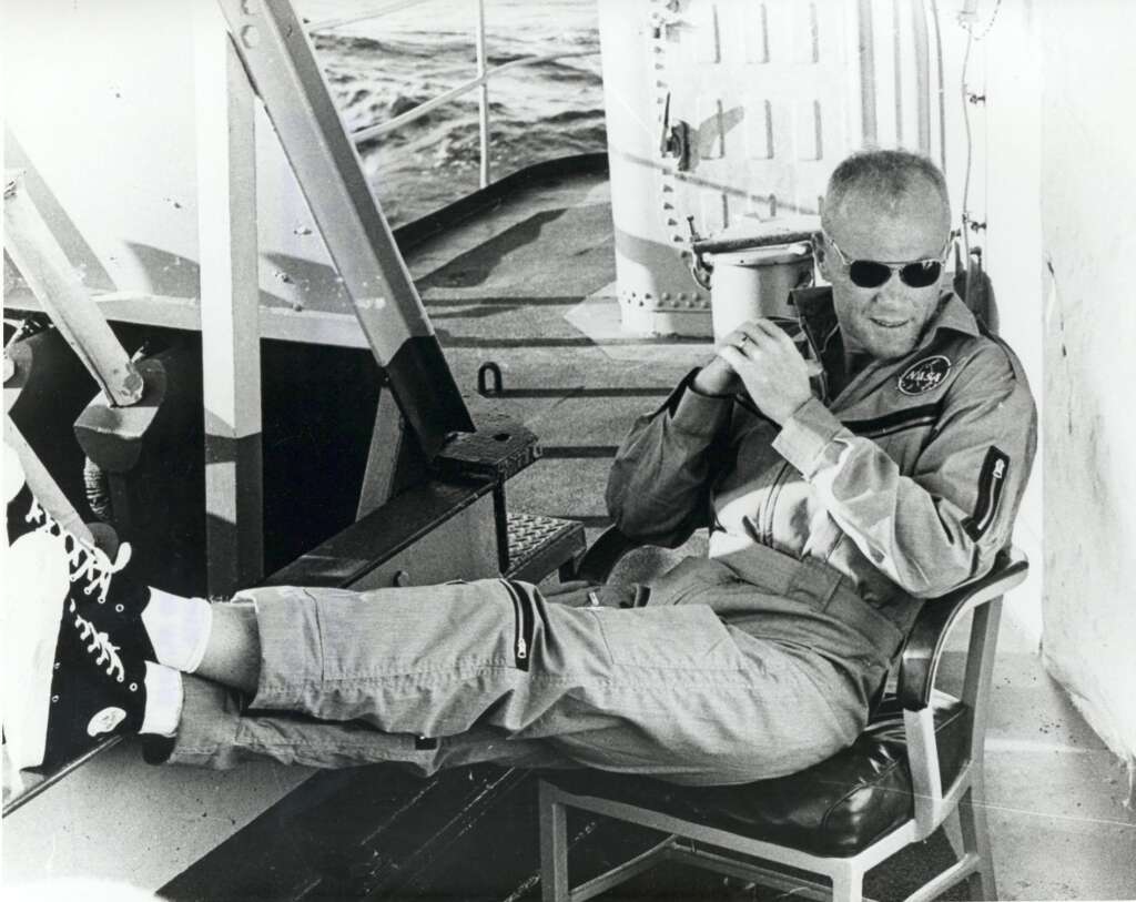 A person wearing a NASA uniform and sunglasses relaxes on a chair aboard a ship, with legs crossed and feet propped up on a railing. They hold a cup, leaning back comfortably. In the background, there's part of the ship and water.