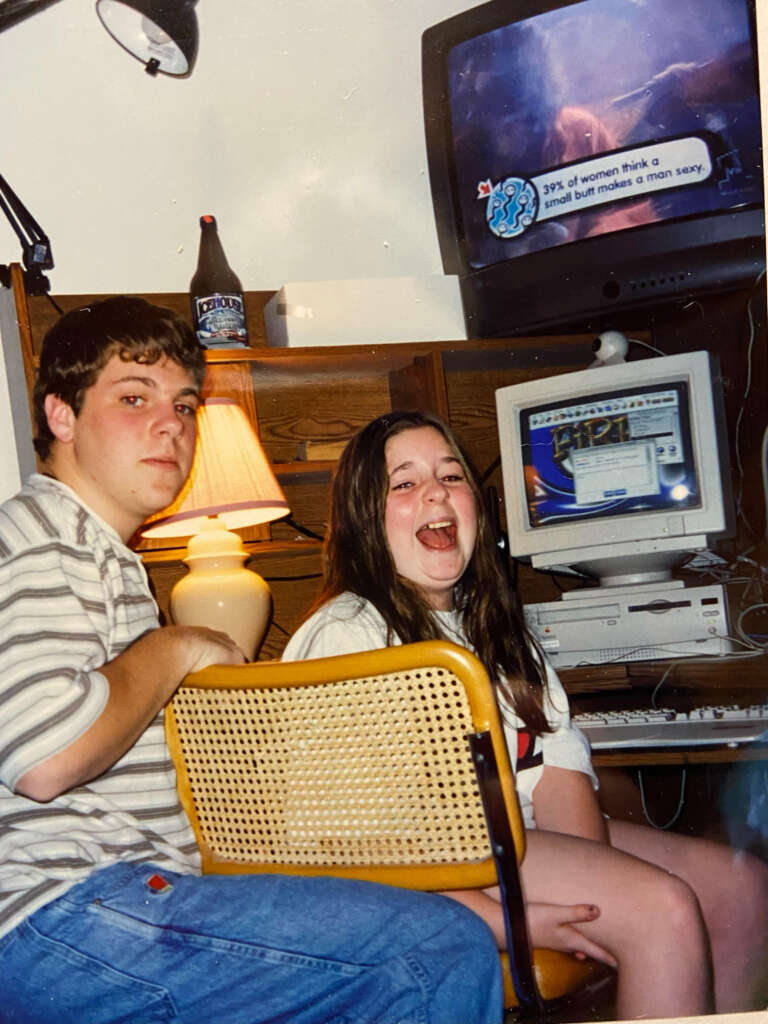 Two people, a young man and woman, are sitting in front of a desk with an old computer setup and a CRT TV on top. They are both looking slightly toward the camera, with the man smiling and the woman laughing. The room has wooden furniture and a table lamp.
