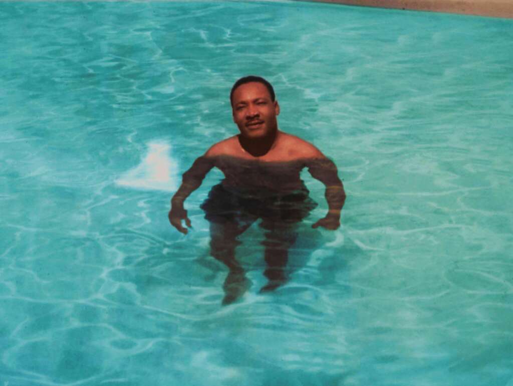 MLK Jr. standing in a swimming pool with water up to his chest. He has a relaxed expression and appears to be enjoying the water. The pool water is clear and blue. The background shows part of the pool’s edge.