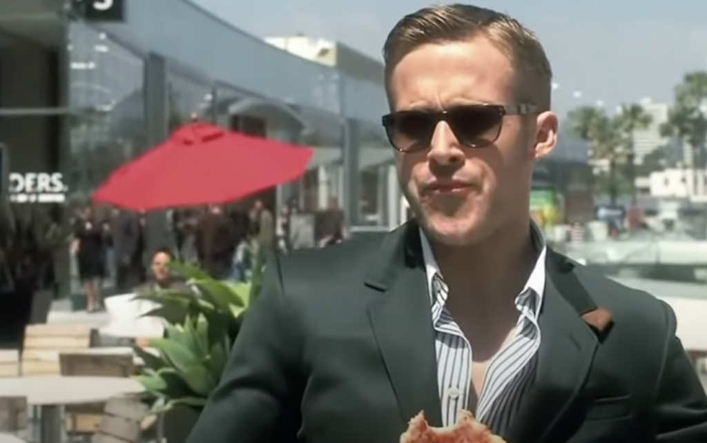 A man with short hair wearing dark sunglasses and a green suit jacket is eating a slice of pizza outdoors. There are people in the background, along with a building, a red umbrella, and some greenery. The man appears to be squinting in the sunlight.