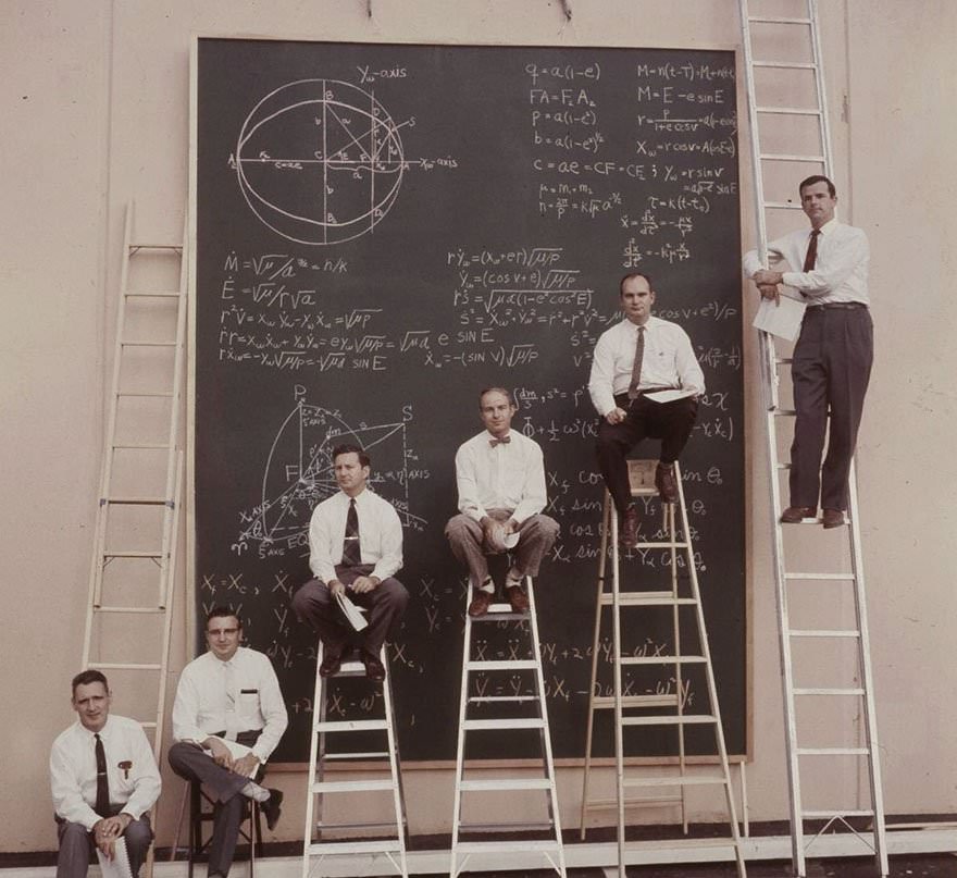 Six men in white shirts and ties are positioned around a large blackboard filled with complex mathematical equations, diagrams, and formulas. Three sit on stepladders, two stand on ladders, and one kneels on the ground.