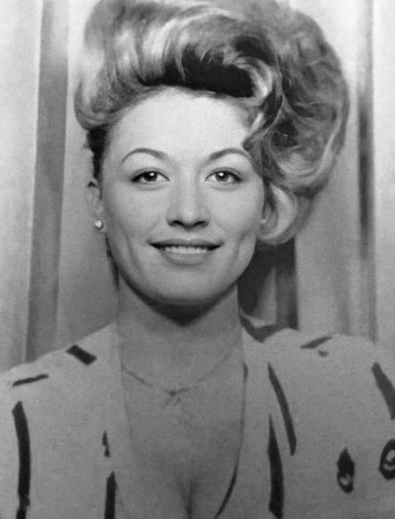 Black and white photo of a woman with styled wavy hair, smiling directly at the camera. She is wearing a light-colored top with a patterned design and earrings. The background features vertical drapes.