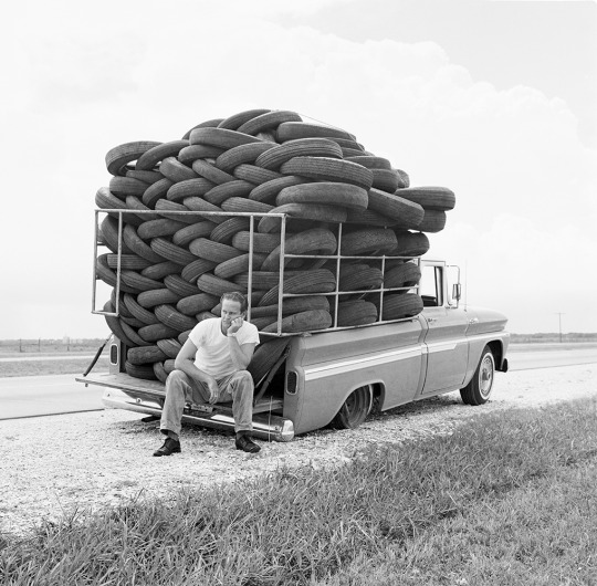 A man sits on the rear of an overloaded pickup truck, which is stacked high with a large, tightly packed collection of tires. The truck appears weighed down, with the rear end near the ground. The scene is set on the side of a rural road with open fields.