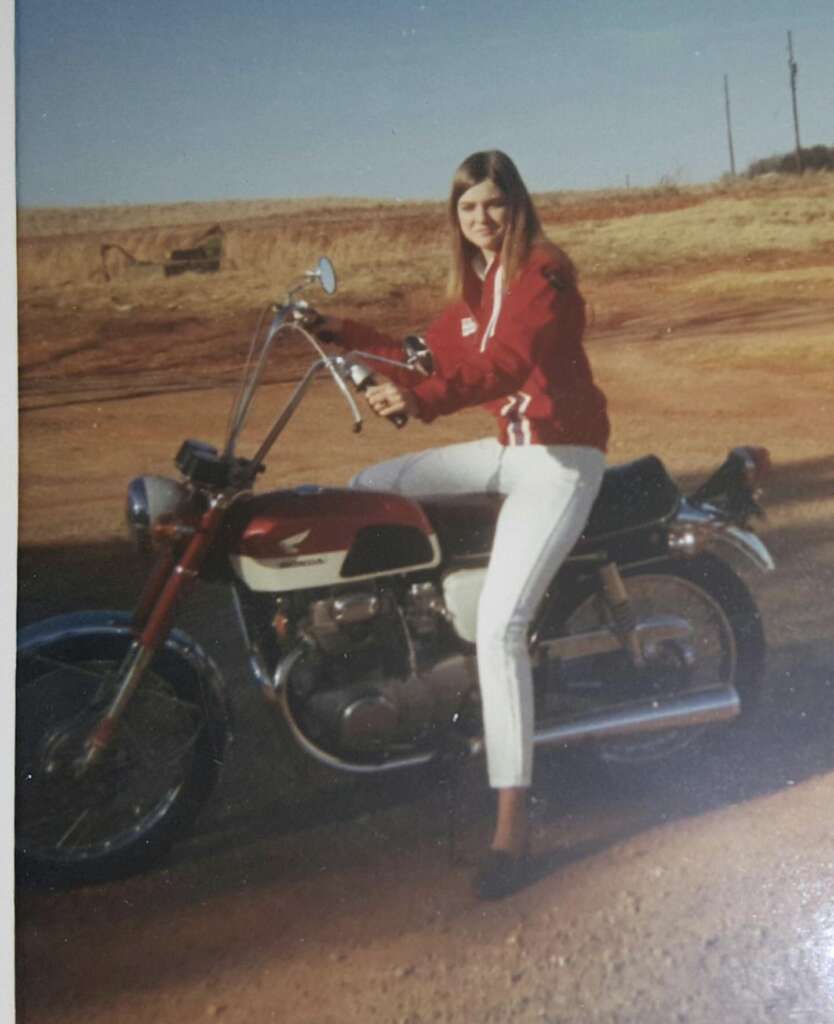 A woman with long hair, dressed in a red jacket and white pants, is sitting on a vintage Honda motorcycle. The background features a dirt road, an open field, and a distant cow. The scene appears to be a sunny day in a rural area.