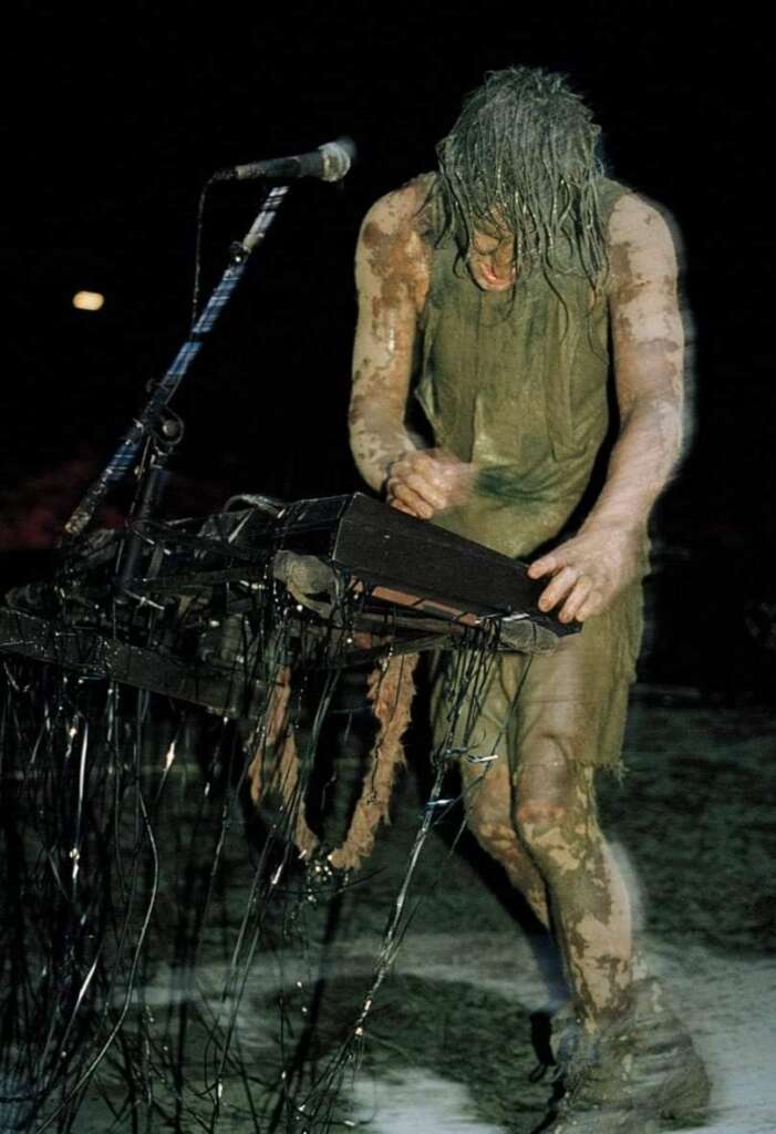 A person stands in front of a microphone and plays a keyboard, covered in mud and with wet hair hanging down over their face. The stage is also muddy and dark, suggesting an intense performance in challenging conditions.