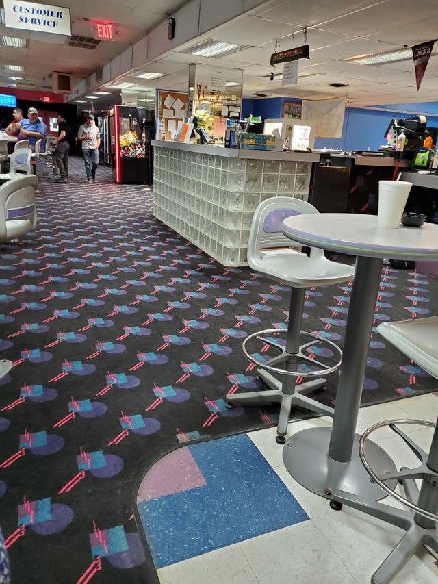 A brightly lit bowling alley with a patterned carpet, featuring a customer service counter, a concession stand with various snacks, and an area with arcade games. In the foreground, a round table with bar stools, one of which has a drink cup on it.
