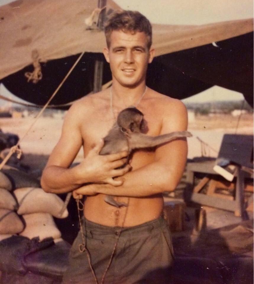 A shirtless man, smiling, holds a small monkey in his arms. The man appears to be outside, standing in front of a tent and various items including a chair and a pile of sandbags. The environment looks like a camp or outdoor site.