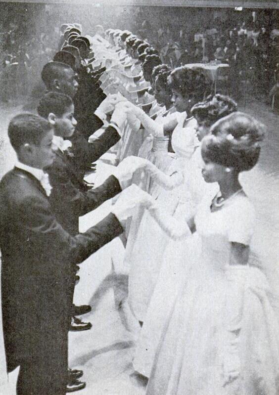 A black-and-white photo depicts a line of young men and women, dressed in formal attire, paired off and facing each other, holding hands. The women are in white gowns while the men wear dark suits and bow ties. They appear to be participating in a formal dance or event.