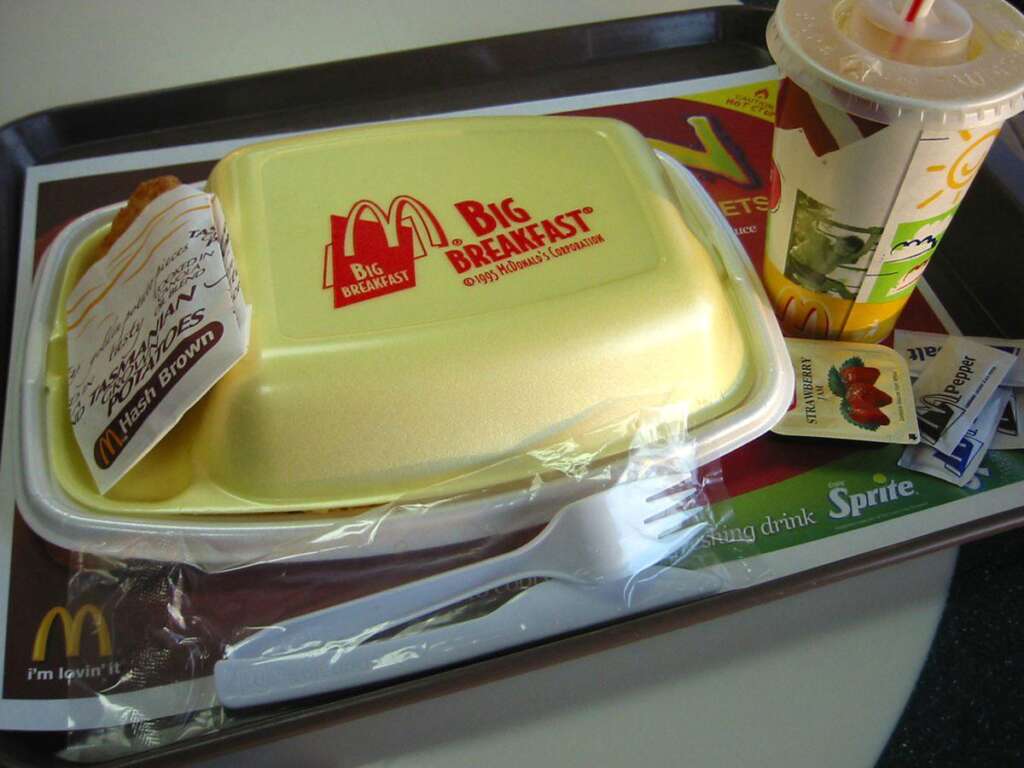 A McDonald's Big Breakfast meal on a tray. The meal includes a yellow-lidded container, a hash brown in a sleeve, utensils in plastic wrap, a drink cup with a straw, and various condiments including a packet of strawberry preserves.