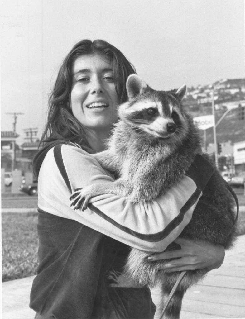 A smiling woman with long hair holds a raccoon in her arms. They are outdoors near a street with buildings and signs visible in the background. The woman is dressed in casual clothing, and the raccoon appears calm and content.