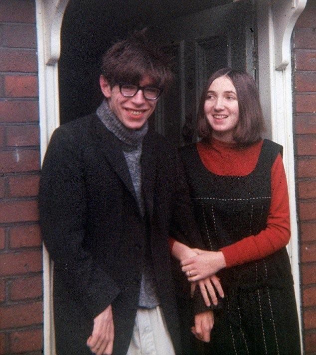 A man with glasses and a dark blazer stands next to a woman dressed in a black dress with a red long-sleeve shirt. They are holding hands and smiling while standing in a doorway. The setting appears to be the entrance of a brick house.