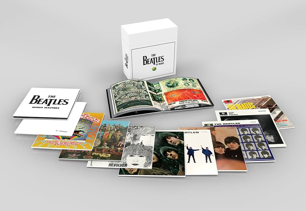 A collection of The Beatles' albums is spread out in front of a white box set labeled "The Beatles." The albums include various iconic covers such as "Sgt. Pepper's Lonely Hearts Club Band," "Revolver," and "Abbey Road." An open booklet shows colorful artwork.