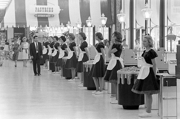 Black and white photo of a grocery store with multiple female cashiers in uniform standing behind registers in a row. The store has a high ceiling and appears spacious. A sign reading "PASTRIES" is visible in the background and there are shoppers in the distance.