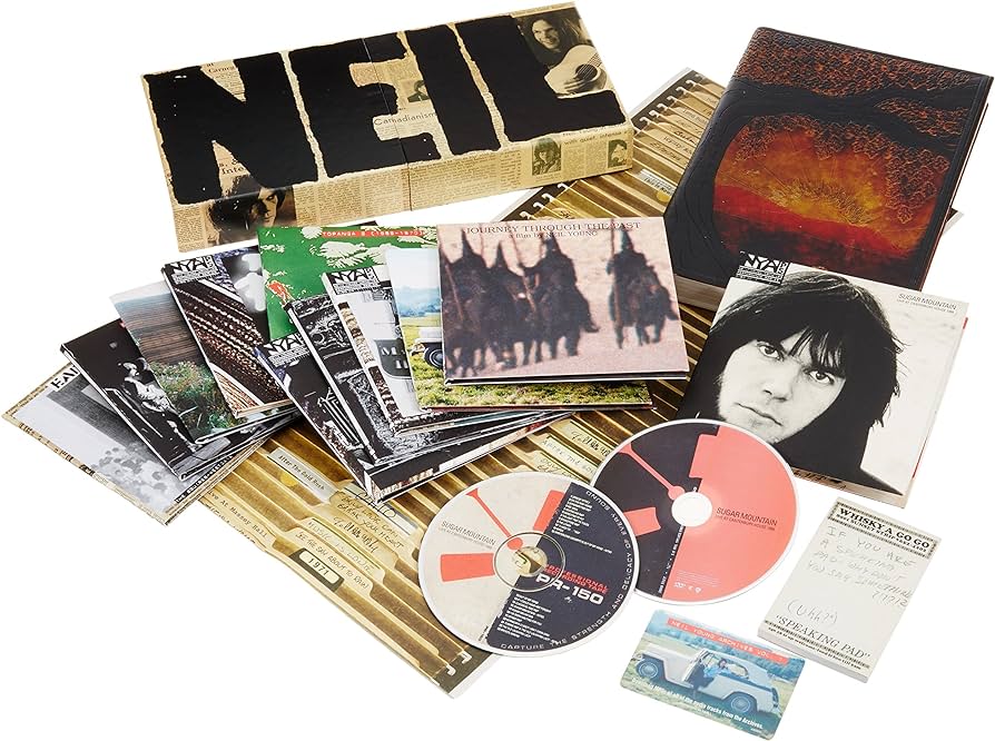 A collection of music albums, CDs, and ephemera spread out on a surface. A large item in the background spells out "NEIL" with a black and white photo. The collection includes various album covers, photos, and CDs with distinct cover art and designs.