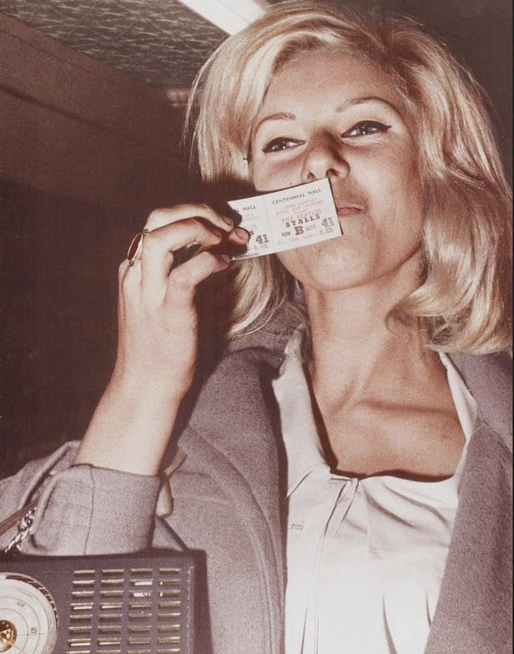 A woman with blonde hair holds a ticket close to her face, partially obscuring her mouth. She is wearing a light-colored top and a coat, and she has a radio or similar device strapped to her shoulder. The image has a vintage feel.