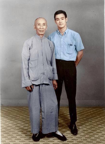 A vintage photo showing two men standing side by side. The older man on the left wears traditional loose-fitting, light-colored Chinese attire, while the younger man on the right wears a short-sleeve, button-up shirt with dark trousers. They are posing against a plain backdrop.