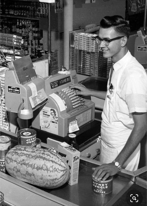 A smiling male cashier, wearing glasses and a white uniform, stands behind an old-fashioned cash register in a grocery store. On the counter are a large watermelon, a can of Jitney Jungle Coffee, two soup cans, and other items.