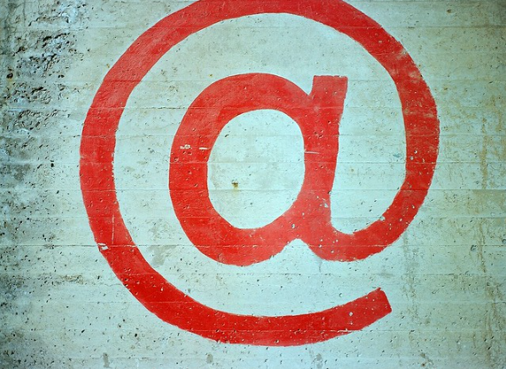 A red at symbol (@) is painted on a textured, light colored concrete wall. The symbol is centered and stands out prominently against the rough surface.