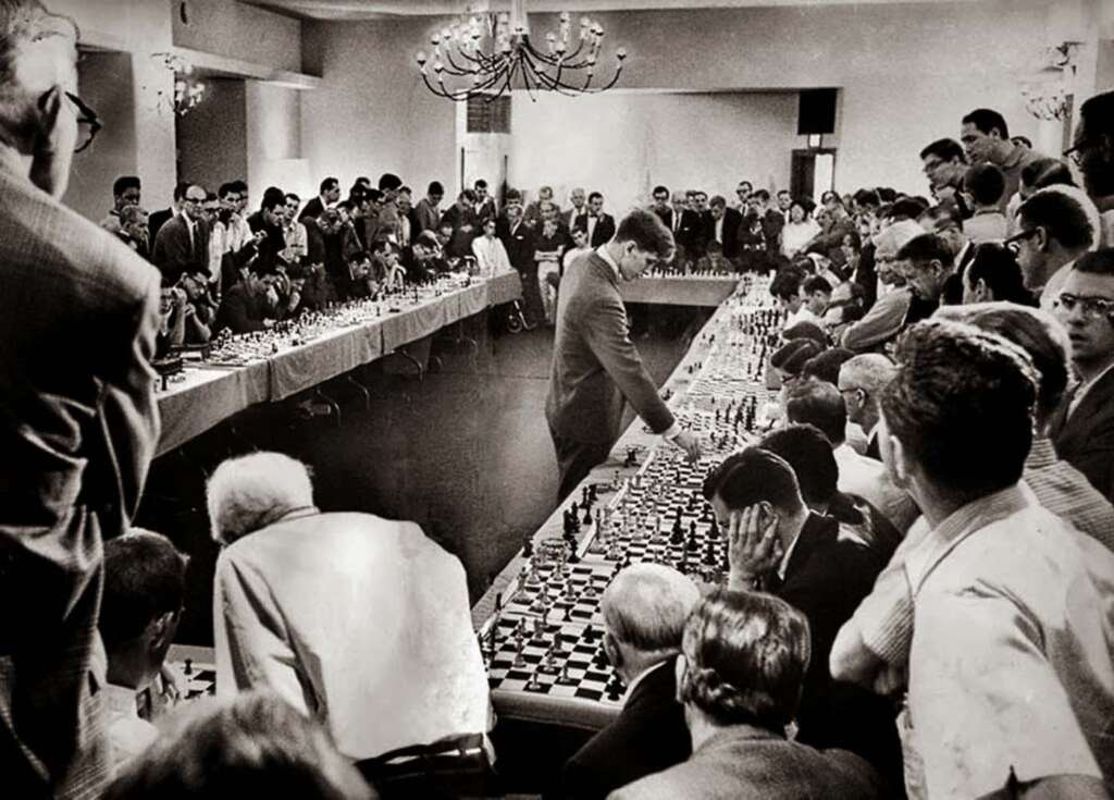 A man is playing multiple games of chess simultaneously in a large, crowded hall. Numerous spectators and players are gathered around several rows of chess tables, observing the intense match. The room is filled with focused attention and strategic gameplay.