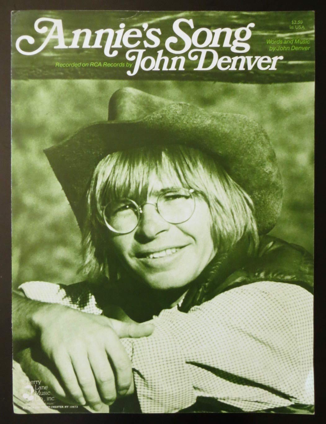 A green-tinted image features a person with long hair, glasses, and a cowboy hat smiling at the camera. The text at the top reads "Annie's Song by John Denver" and mentions it's recorded on RCA Records. Additional text credits John Denver for words and music.