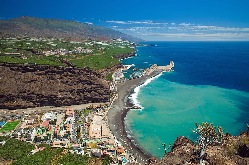 A scenic view of a coastal town with a mix of buildings, beaches, and green fields. The shoreline curves, leading to a harbor with boats, while the turquoise ocean contrasts with the rocky cliffs and mountains in the background under a clear blue sky.