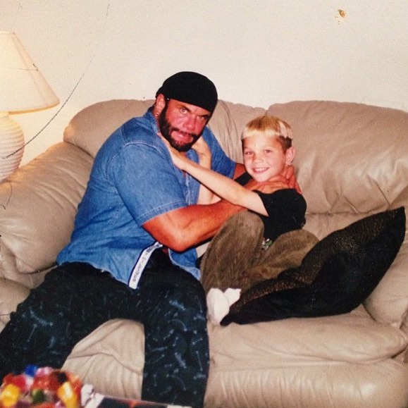 A man wearing a denim shirt, black beanie, and black pants playfully embraces a young boy on a beige sofa. The boy, smiling broadly, wears a light-colored shirt and dark pants. The background includes a white lamp and a part of a table with some objects.