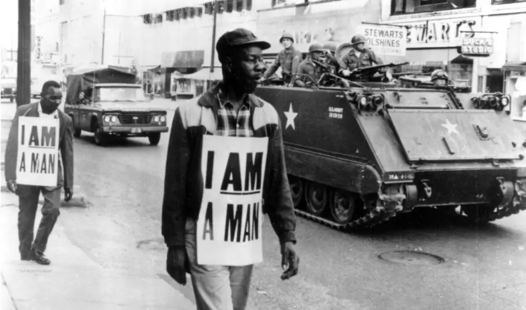 Black and white photo of two men marching in the street wearing placards that read "I Am A Man." In the background, there is an armored military vehicle and several soldiers standing atop it. Additional onlookers and buildings are visible in the background.