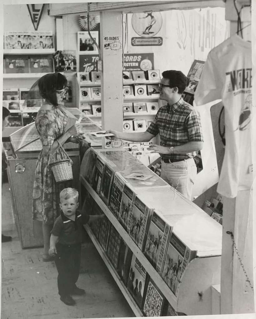 Black and white photo showing a woman and a child at a record store counter. The woman is in a patterned dress, the child looks towards the camera, and the clerk is a man in glasses and a plaid shirt. The store has shelves of records and some hanging clothing.