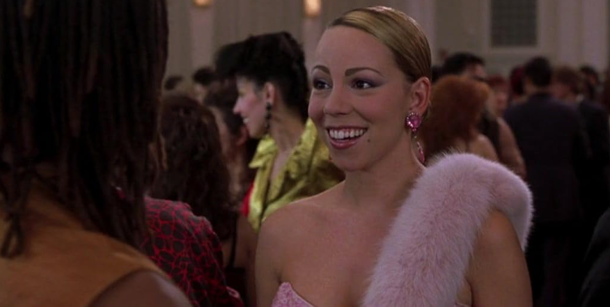 A woman with a bright smile is dressed in a glamorous pink outfit and earrings, standing in a crowded party scene. She is looking at another person in the foreground whose face is not visible. The background is blurred, with other attendees in fashionable attire.