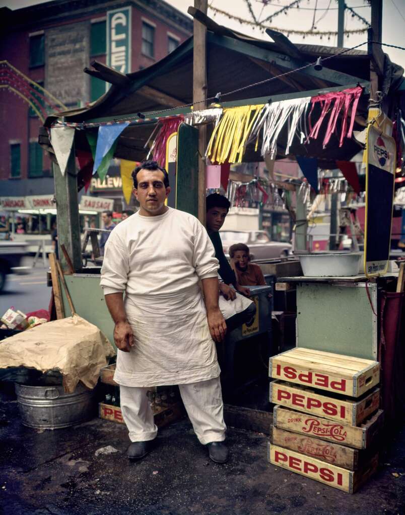 A man in a white apron and white attire stands in front of a makeshift food stall decorated with colorful bunting. Behind him, a person is seated and another is partially visible. Crates of Pepsi surround the stall. The background shows a busy street and buildings.