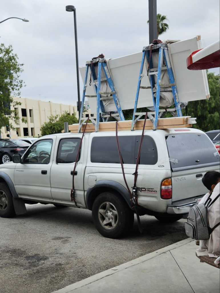 A white Toyota pickup truck is parked on the street with large wooden pieces and ladders secured to its roof with red straps. The truck has a camper shell, and part of a person wearing a patterned jacket is visible in the bottom right corner of the frame.