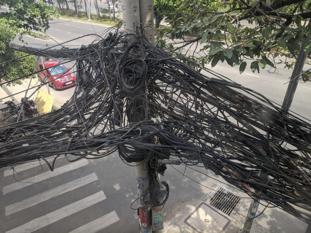 A utility pole is cluttered with a mass of tangled wires, creating a chaotic appearance. The pole is situated near a crosswalk and a road with light traffic. A red car is parked nearby, and trees with green foliage are visible along the street.