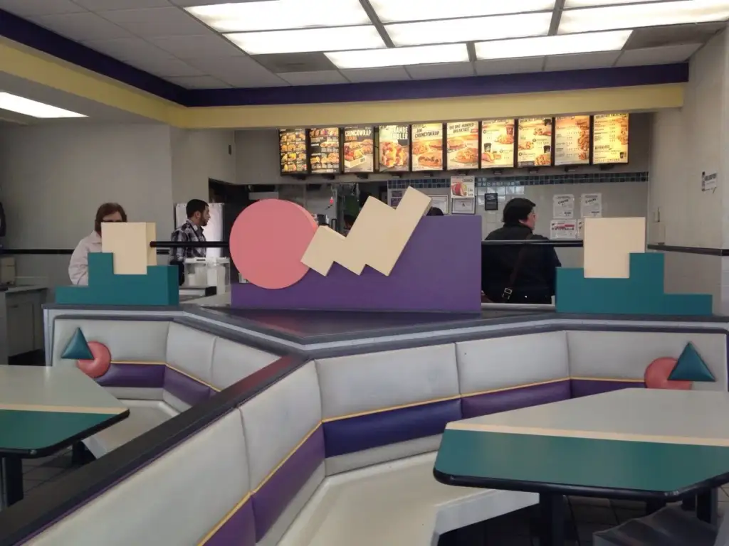 A fast-food restaurant interior with booth seating featuring colorful geometric shapes. The back wall has a counter, several menu boards, and employees attending to customers. The overall decor includes various pastel colors and bold shapes, giving it a retro feel.