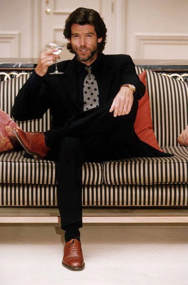 A man with dark hair and a beard sits on a striped couch, holding a glass of wine. He is wearing a black suit with a grey polka-dot tie and brown shoes. There are red pillows on the couch and a light-colored wall in the background. The man is smiling slightly.