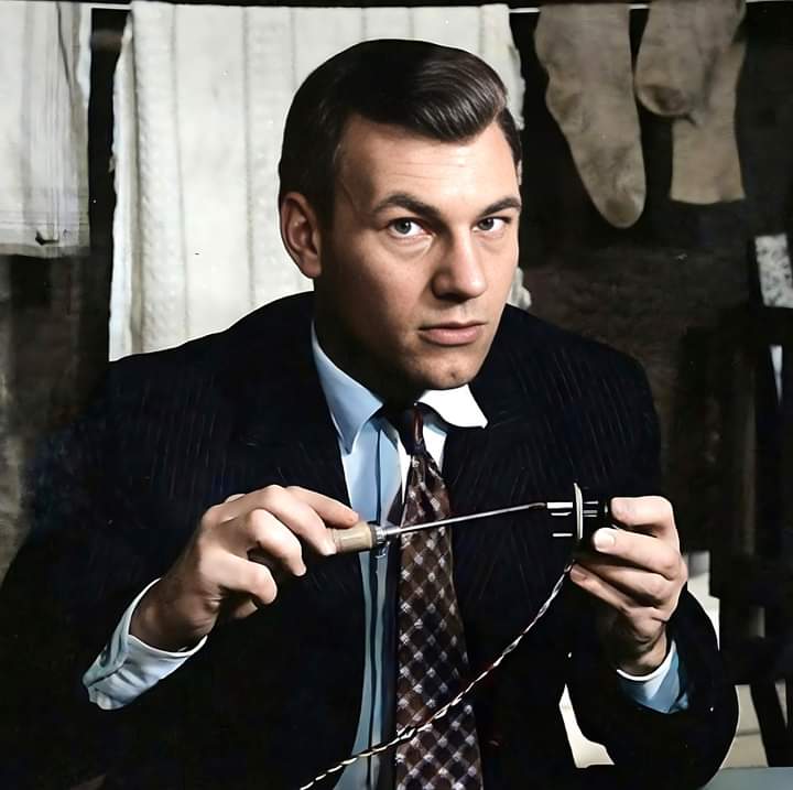 A man in a suit and tie is working with an electronic device, holding a tool and what appears to be a spool or component. There is a clothesline with hanging laundry in the background. He has a serious expression and is positioned in an indoor setting.