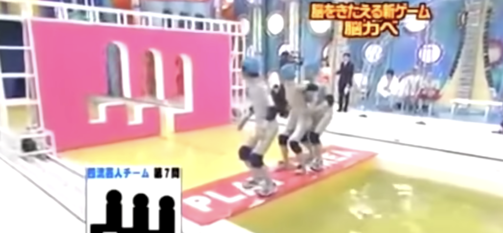 Four people wearing silver suits and helmets are trying to form a specific shape to pass through a cut-out wall on a Japanese game show. They are standing on a red platform labeled "PLAY" by a pool of water, with a challenging hole shape approaching them.