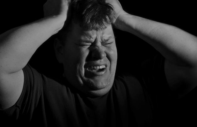 A man with his eyes closed and face contorted in pain or frustration clutches his head with both hands. The background is dark, highlighting his expression and emphasizing his emotional state.