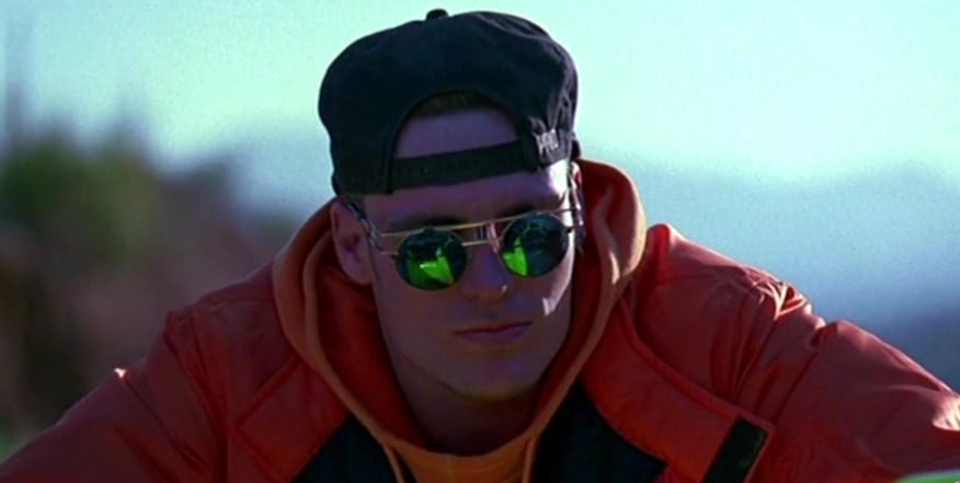 A person wearing a backward black cap, mirrored sunglasses, and a red jacket looks off to the side. The reflective sunglasses show a green reflection, and the background hints at an outdoor setting with blurred elements.