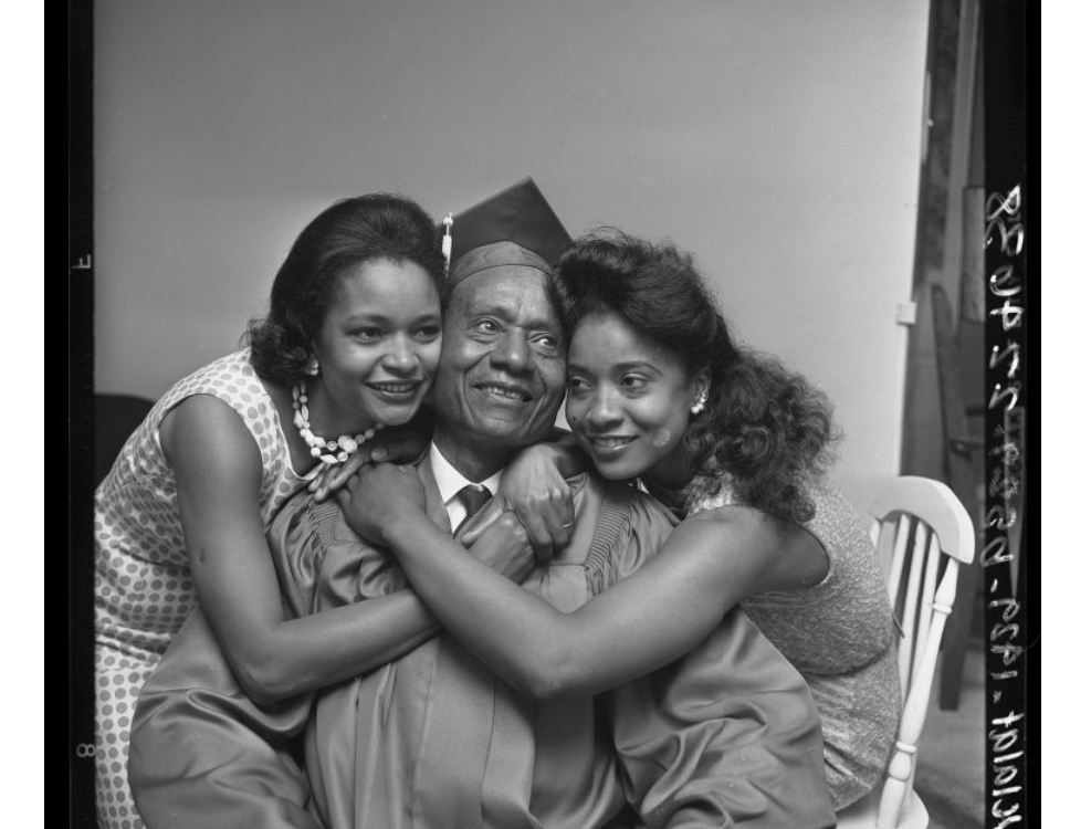 A black and white photo shows a graduate, wearing a cap and gown, being hugged by two women on either side. They all smile joyfully. The background is plain and a white chair is partially visible behind the woman on the right.