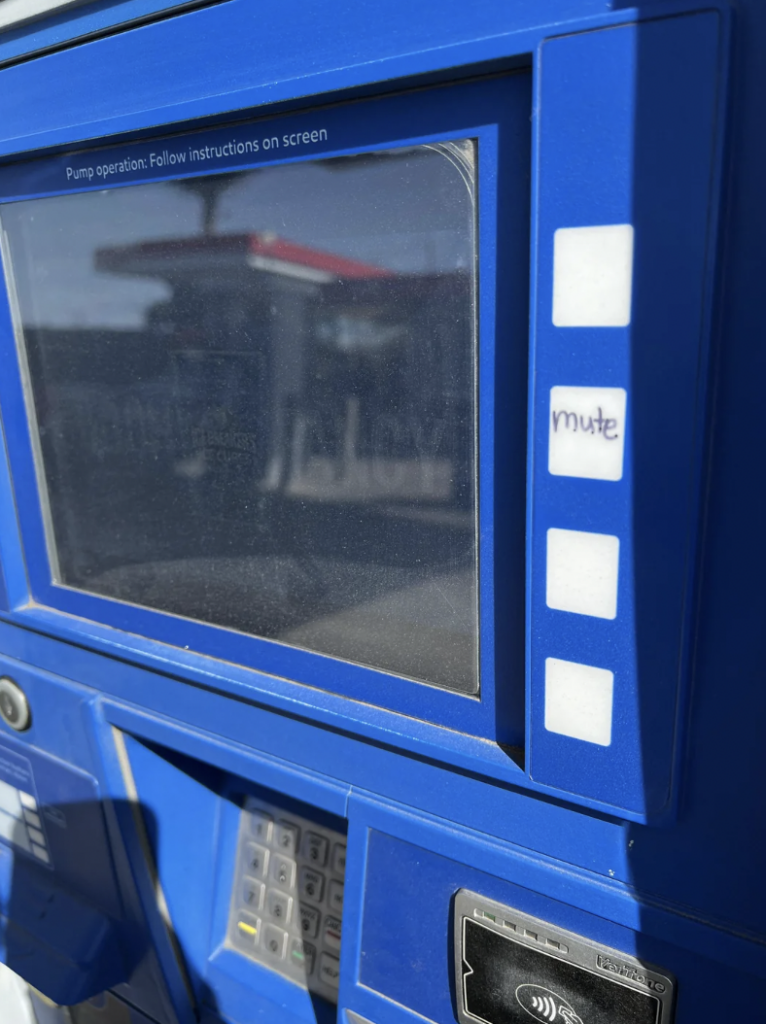 A close-up view of a blue gas station pump screen with a label next to one of the buttons that reads "mute." The screen is turned off and instructions are written above it. A card reader and keypad are visible below the screen.