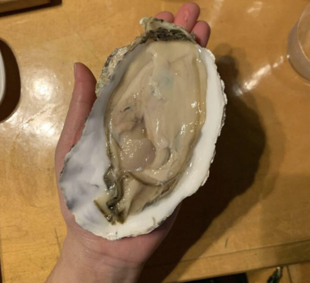 A hand holding a large, opened oyster on the half shell, displaying its natural, moist interior. The shell has a rugged, textured exterior, and the background consists of a smooth, light brown surface.