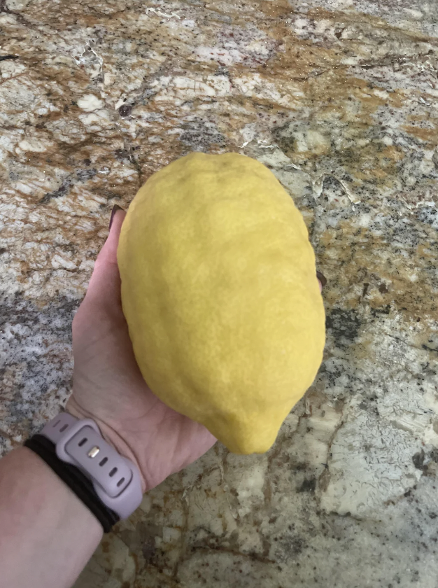 A person with a smartwatch on their wrist holds an unusually large, bright yellow lemon against a marble countertop.