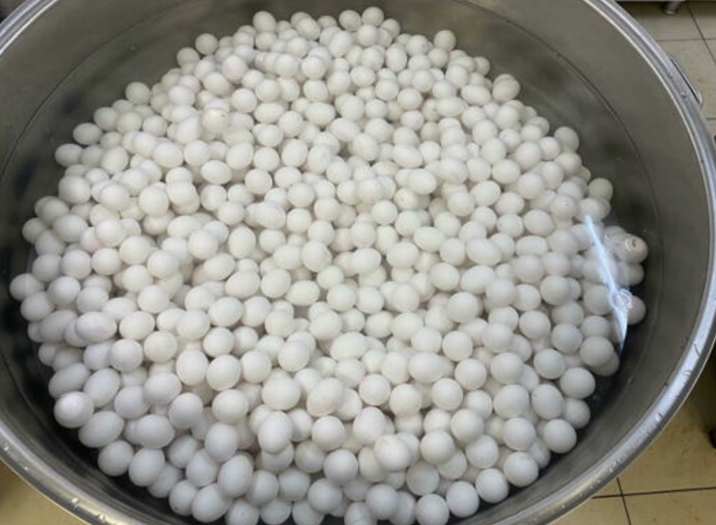 A large container filled to the brim with numerous white chicken eggs. The eggs are clean and uniformly shaped. The container has metallic sides, and a small section of the container's edge is visible. The background is a tiled surface.