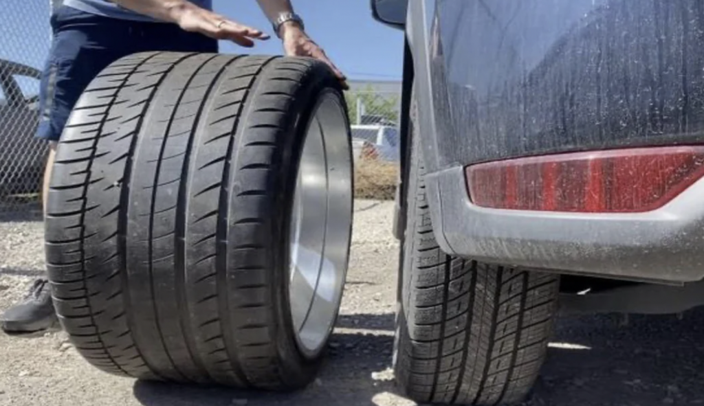 A person comparing the size of a large, wide tire placed vertically on the ground with a smaller tire mounted on the back of a vehicle. The scene takes place outdoors, with a chain-link fence in the background.