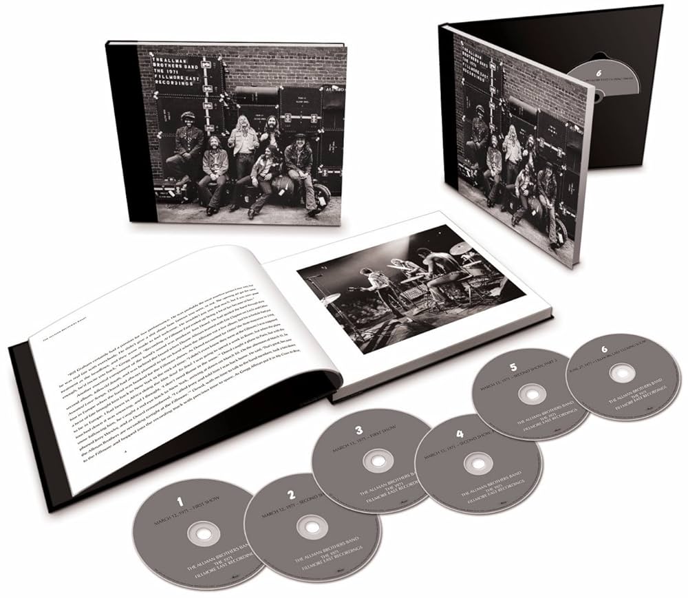 A music box set is displayed, featuring a book with band photos and text, along with six CDs labeled from 1 to 6. One CD case is open, showcasing the same band photo as the book. The entire set is arranged neatly on a white surface.