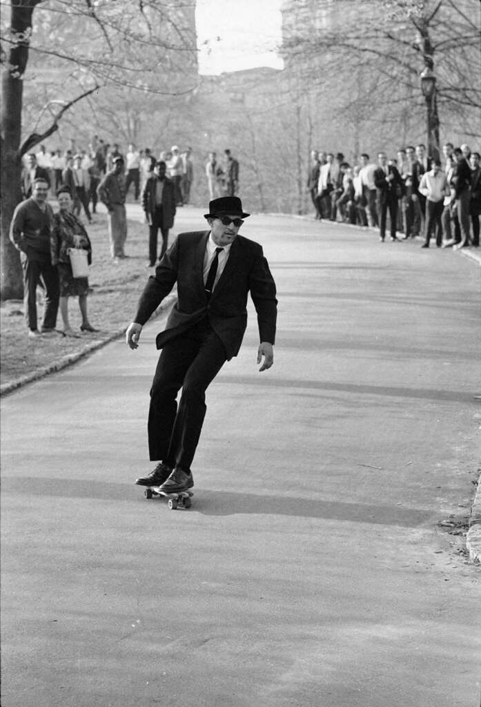 A man in a suit and hat skateboards down a park pathway. He is balancing confidently while the skateboard tilts to the left. In the background, a crowd of people stands and sits on the grass, watching him. Trees line the path, and it appears to be a sunny day.