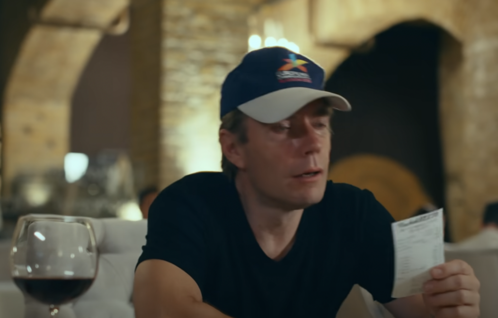 A person wearing a blue cap is sitting at a table in a restaurant, holding a piece of paper, possibly a receipt, in one hand. There is a glass of red wine in front of them. The background shows a rustic interior with stone walls and arches.