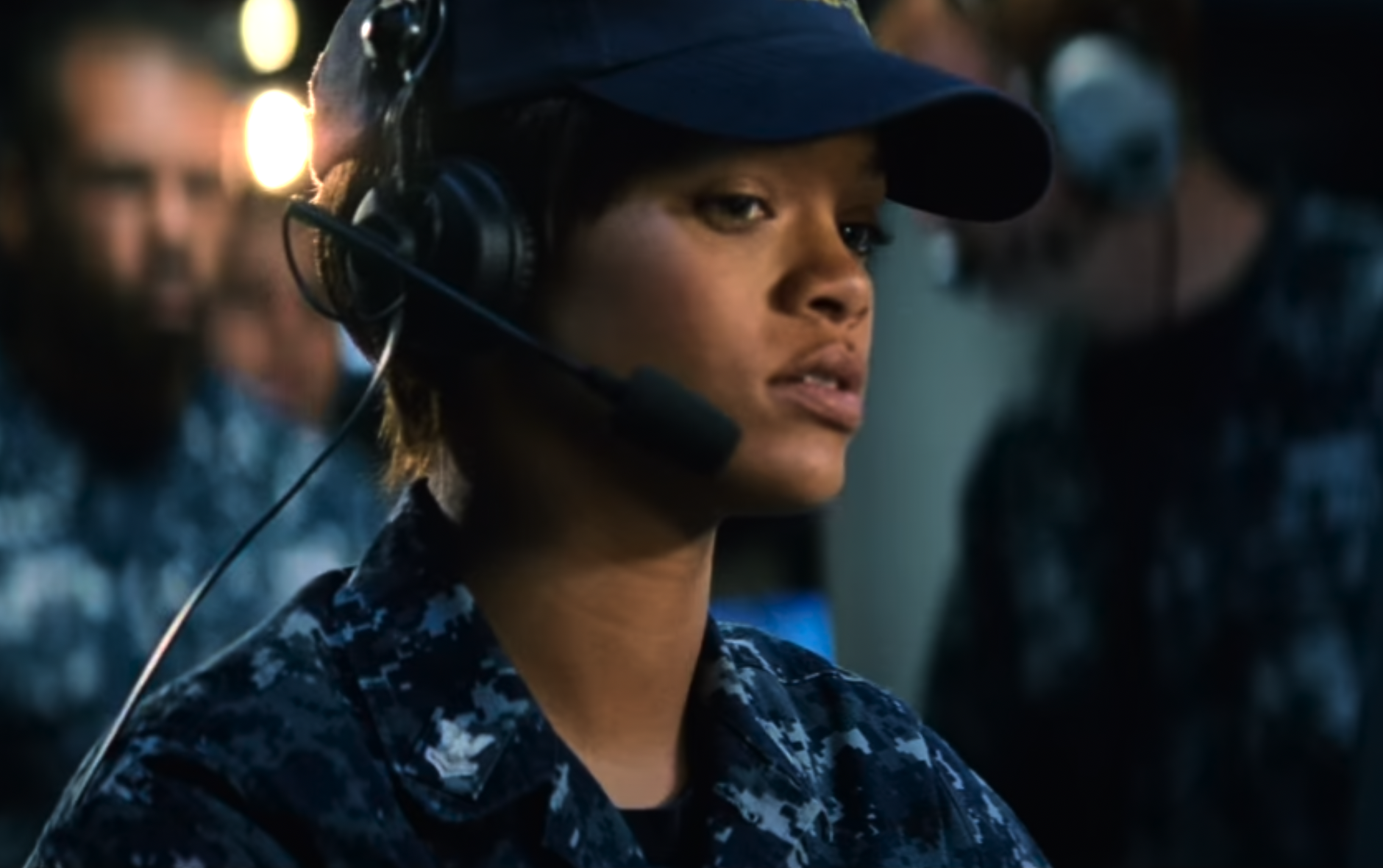 A person in military attire, wearing a camouflage uniform and a cap, is shown with a headset and microphone. They appear focused and engaged in some activity. Several other individuals in similar attire are visible in the blurred background.