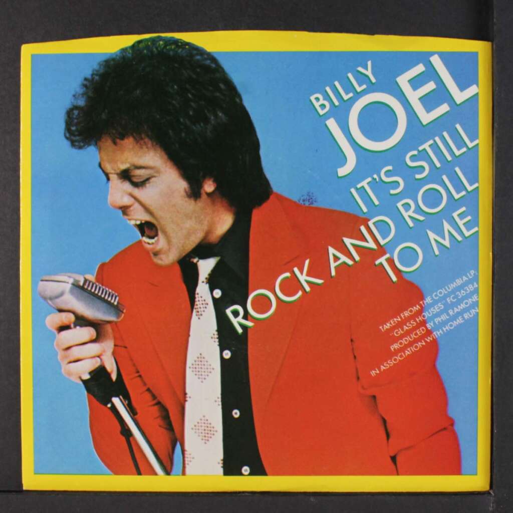 A man with black hair, wearing a red jacket and white polka-dotted black tie, passionately sings into a vintage microphone on the cover of an album. The text reads "Billy Joel - It's Still Rock and Roll To Me." The background is bright blue and yellow.